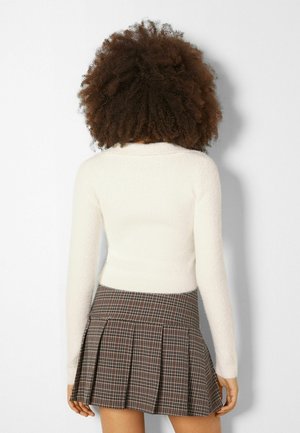 Femme aux cheveux bouclés portant un haut blanc à manches longues et une jupe plissée à carreaux marron, vue de dos sur un fond uni.