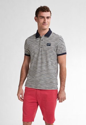 Streifen-Polohemd mit marineblauem Kragen und kurzen Ärmeln; der Stoff hat eine strukturierte Oberfläche. Kombiniert mit roten Shorts. Kleines Logo auf der Brust.