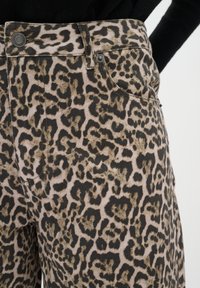 Gros plan sur un pantalon à imprimé léopard avec poches avant et fermeture par bouton métallique, porté avec un haut noir.