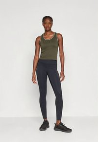 Leggings pretas combinadas com uma camisola de alças verde-oliva com uma borda branca. A modelo está de pé, calçando ténis desportivos pretos, num fundo simples.
