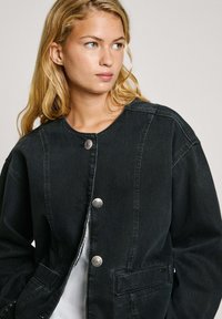 Schwarze Denimjacke mit rundem Ausschnitt, silbernen Druckknöpfen, zwei Fronttaschen und lockerer Passform. Verfügt über einen strukturierten Stoff.