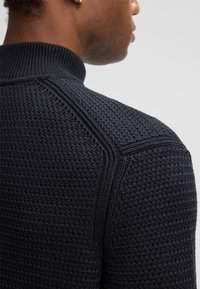 Donkerblauwe gebreide trui met een hoge kraag, een gestructureerd zigzagpatroon en onderscheidende schoudernaaddetails.