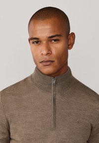 Bruine ritssweater met hoge kraag, gemaakt van soepele stof, met een klein logo op de borst en een getailleerd ontwerp.