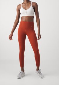 Kobieta w białym staniku sportowym, w wysokich, pomarańczowych legginsach z kieszeniami po bokach oraz jasnoszarych sneakersach Nike, stojąca na jednolitym tle.