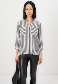 Grijze en witte gestreepte blouse met een V-hals, twee voorzakken en driekwart mouwen, gecombineerd met zwarte strakke broeken.