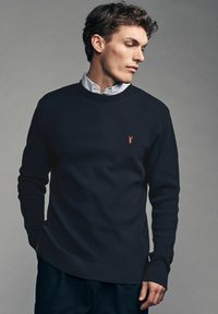 Tumedast merevaigist tehtud crewneck kampsun tekstuurse kudumisega mustris, millel on rinnal väike punane homaarieemblem. Pikad varrukad, soonikkoes mansetid ja alläär.