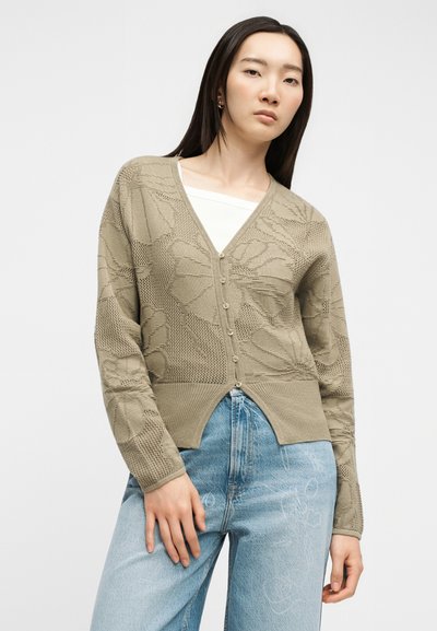Mujer que lleva una chaqueta de punto beige con botones y escote en V con patrón floral, camiseta interior blanca y vaqueros azul claro, de pie contra un fondo liso.