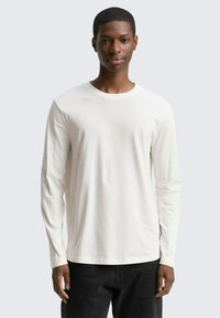 Lange mouwen shirt in licht beige stof, ronde hals, relaxte pasvorm, gladde textuur, gedragen met zwarte broek, model kijkt recht vooruit.