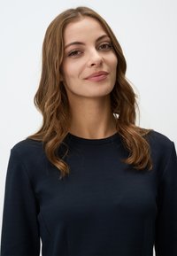 Top à manches longues bleu marine avec un col rond, texture lisse et design ajusté. Cheveux ondulés tombant sous les épaules.