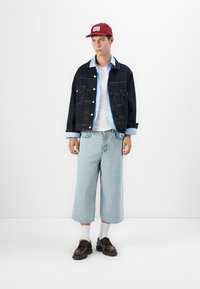 Denimjack met blauwe accenten, witte t-shirt, lichtblauwe baggy jeans, rode pet, witte sokken en bruine veterschoenen, tegen een effen achtergrond.