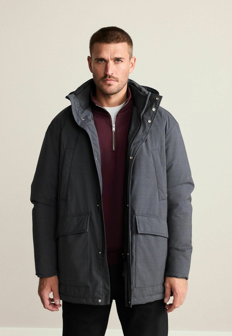 Mann mit kurzen Haaren, der eine zweifarbige schwarze und graue Winterjacke über einem kastanienroten Reißverschlussoberteil trägt, steht vor einem schlichten hellen Hintergrund.