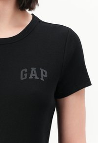 Primo piano di una persona che indossa una maglietta nera a coste con maniche corte e la scritta "GAP" in grigio scuro sul lato sinistro del petto.