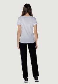 T-shirt bianco a maniche corte, design aderente, abbinato a pantaloni neri a gamba dritta. Dettaglio visibile nella schiena e calzature casual in scala di grigi.