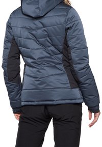 Frau trägt eine blaue gepolsterte Jacke mit schwarzen Seitenpaneelen und Kapuze, steht mit dem Rücken zur Kamera vor einem weißen Hintergrund.