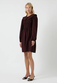 Abito bordeaux con corpino arricciato, maniche lunghe a palloncino e un grande colletto con un motivo a quadretti. Completano il look delle mule nere con tacco.