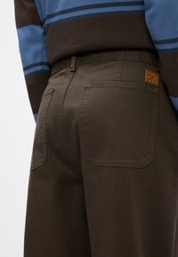 Brune bomullsbukser med en jevn tekstur, med to baklommer og en liten logo-patch i brunt skinn.