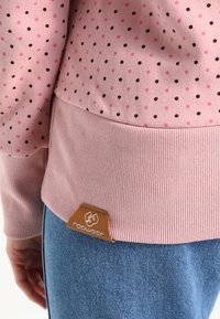 Pull rose à petits pois roses et noirs, ourlet côtelé avec étiquette logo marron, porté avec un jean en denim bleu à la taille d’une personne.