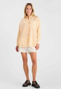 Femme portant une chemise à rayures jaunes et blanches, ample et boutonnée, un short texturé blanc et des sandales noires à semelles épaisses, se tenant devant un fond uni.