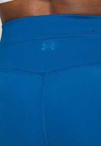 Niebieskie legginsy sportowe z wysokim pasem. Materiał wydaje się gładki, z małym, wytłoczonym logo w dopasowanym odcieniu.