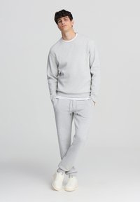 Giovane uomo che indossa un felpa grigia chiara e pantaloni della tuta abbinati con scarpe bianche, in piedi con le mani in tasca su uno sfondo neutro.