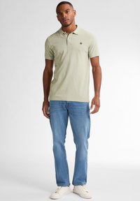 Polo shirt verde chiaro con un piccolo logo, abbinato a jeans blu e sneakers bianche. Il tessuto sembra morbido e traspirante.