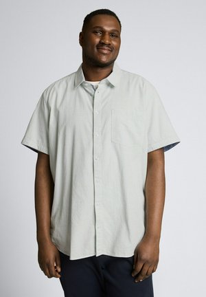 Man smiling, standing, wearing light gray short-sleeve button-up shirt and dark pants against a plain light background.