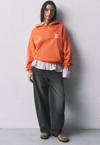 Orange hoodie with front pocket and "Cute But Wild" text, layered over a striped shirt. Pair of loose black jeans and tan footwear.