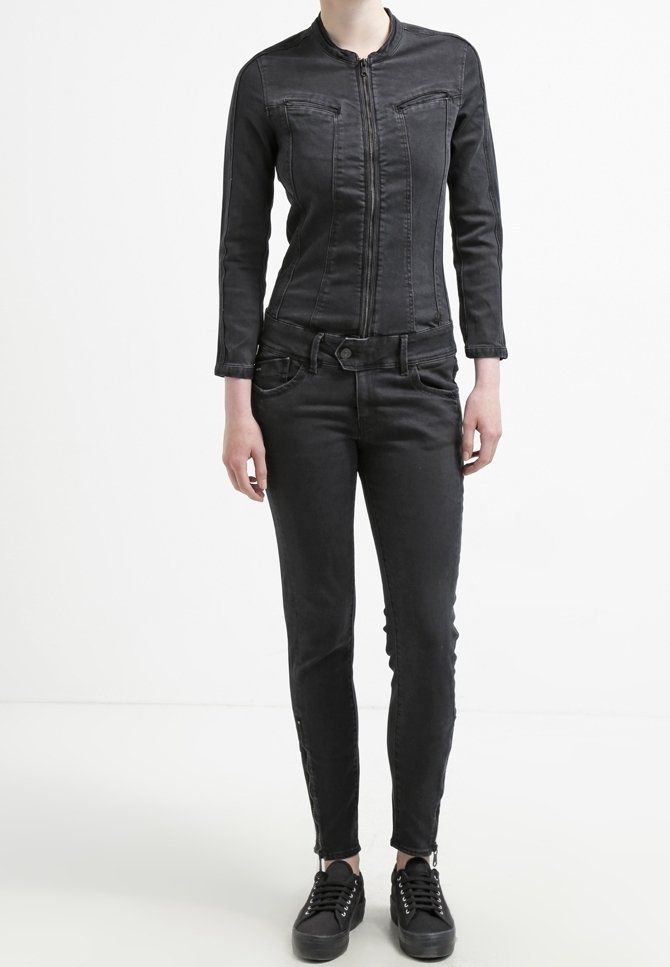 Personne portant une veste noire ajustée à fermeture éclair, un jean noir slim et des baskets noires, debout devant un fond blanc.