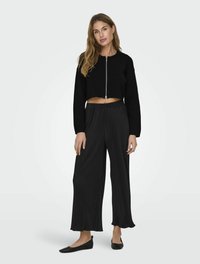 Maglione cropped nero con zip frontale, abbinato a pantaloni neri a gamba larga con una texture a coste e orli svasati; scarpe nere piatte.