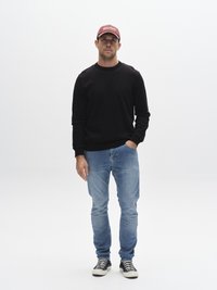 Črni pulover z dolgimi rokavi, svetlo modre jeans hlače in črno-bele črtaste superge. Oseba nosi bordo kapa z belim napisom.