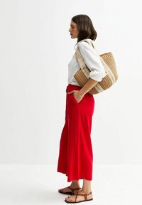Pantalones rojos de pierna ancha, camisa blanca, sandalias de tiras marrones y un bolso de paja tejido con una correa beige. El conjunto presenta una silueta relajada.