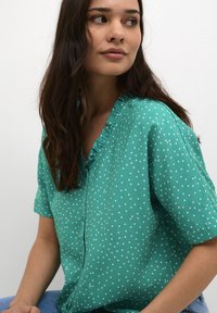 Blusa turchese a maniche corte con pois bianchi, caratterizzata da un colletto arricciato e una chiusura con bottoni. Tessuto morbido con una vestibilità comoda.