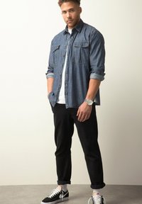 Denim button-up overhemd met twee borstzakken, opgerolde mouwen, gedragen over een wit T-shirt; gecombineerd met zwarte tapered broek en zwarte sneakers.