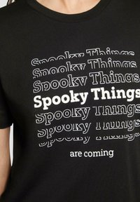 T-shirt en coton noir avec un texte empilé "Choses Effrayantes" en blanc et gris clair, avec des opacités variées et une police en gras soulignant le message.