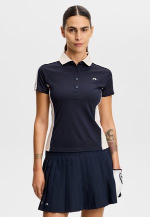 Kobieta ubrana w granatowo-białą krótkorękawową koszulkę polo do golfa i plisowaną spódnicę, z rękawiczką golfową na lewej dłoni oraz widocznymi tatuażami na ramieniu.