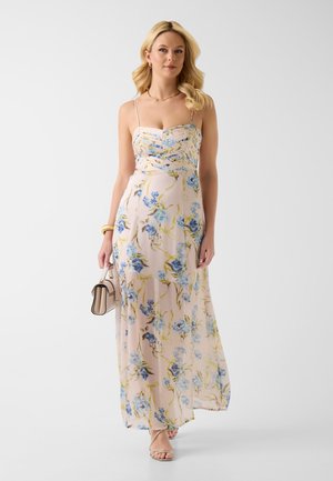 Kobieta w różowej, kwiecistej sukni maxi bez rękawów, trzymająca beżową torebkę, z rozpuszczonym blond włosami, idąca do przodu na jednolitym tle.