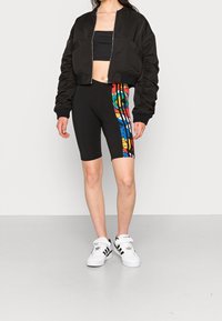 Svart bomberjacka med puffärmar, svart crop top, svarta cykelshorts med en färgglad grafisk design och vita Adidas-sneakers.