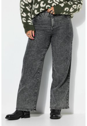 Person iført højttaljede, vidde, falmede sorte jeans, sorte ankelstøvler og en grøn- og hvidmønstret sweater, hånd hvilende på hoften.