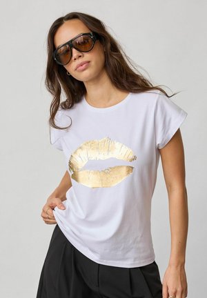 Mujer con gafas de sol negras y una camiseta blanca con una gran impresión de labios en foil dorado, posando con las manos en las caderas.