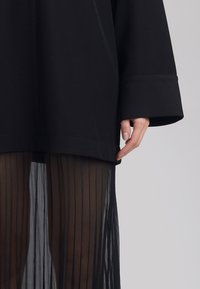 Fekete oversize felső széles ujjakkal, áttetsző fekete redőzött szoknyával párosítva. A felső egyenes szegélyű, a szoknyán függőleges vonalak találhatók.