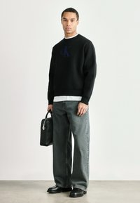 Čierna mikina s modrým logom "CK", skombinovaná so svetlo šedými širokými nohavicami a čiernymi koženými topánkami. Drží obdĺžnikovú čiernu tašku.