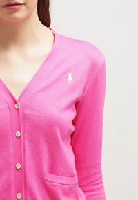 Kvinna i en knallrosa knäppt cardigan med en liten vit polo-spelare-logotyp på vänster bröst.