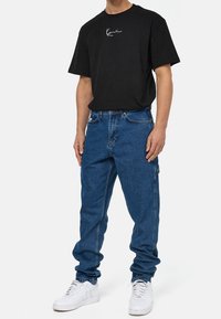 Camiseta negra de manga corta con logo blanco, combinada con jeans cargo azules y zapatillas deportivas blancas. El atuendo tiene un ajuste casual y relajado.