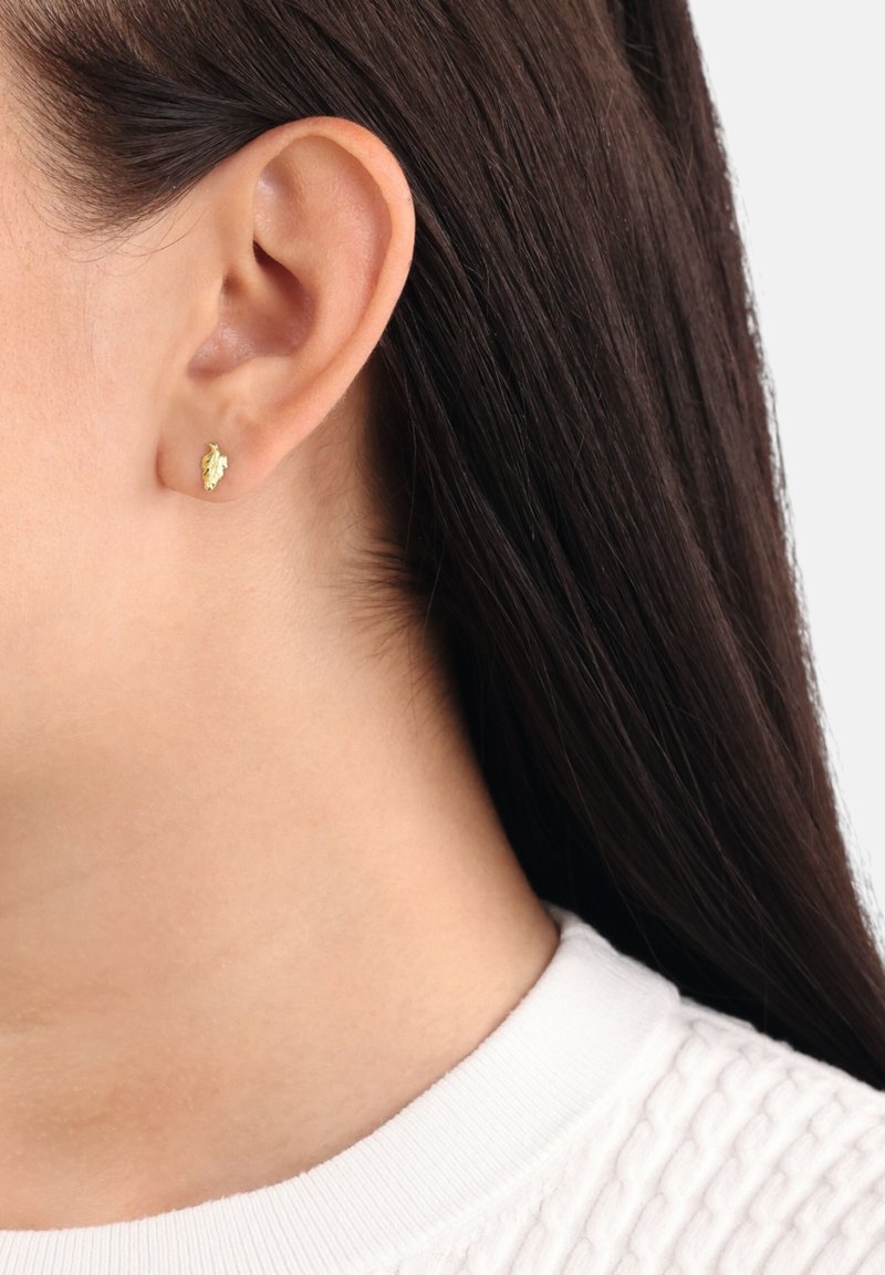 Pendiente de stud en forma de hoja de oro en la oreja de un modelo. Textura suave, tamaño pequeño y diseño minimalista, contrastando con el cabello oscuro y un suéter blanco.