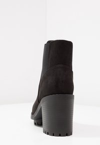 Schwarze Wildleder-Ankle Boots mit klobigem Absatz und elastischem Seiteneinsatz, fotografiert vor einem weißen Hintergrund.