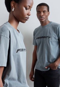 Las camisetas de algodón grises presentan un destacado logotipo negro de "PRIORITY", con cuellos redondos y mangas cortas, llevadas por dos modelos frente a un fondo neutro.