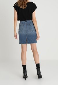 Falda de mezclilla en azul medio con un corte ajustado, detalle de abertura en la parte trasera y bolsillos traseros. Combinada con una blusa negra y botas de tacón alto.
