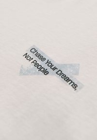 Texte sur un tissu clair : « Poursuis tes rêves, pas les gens » avec deux morceaux de ruban adhésif translucide formant un X derrière le texte.