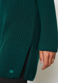 Nahaufnahme eines türkisfarbenen, gerippten Strickpulli-Ärmels mit seitlichem Schlitz, der eine Hand und ein kleines rechteckiges genähtes Etikett nahe dem Saum zeigt.