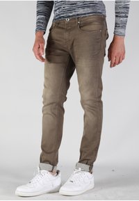 Jeans de mezcla de algodón marrones con corte ajustado, que incluyen bolsillos frontales, cierre de botón y puños doblados, combinados con zapatillas blancas.